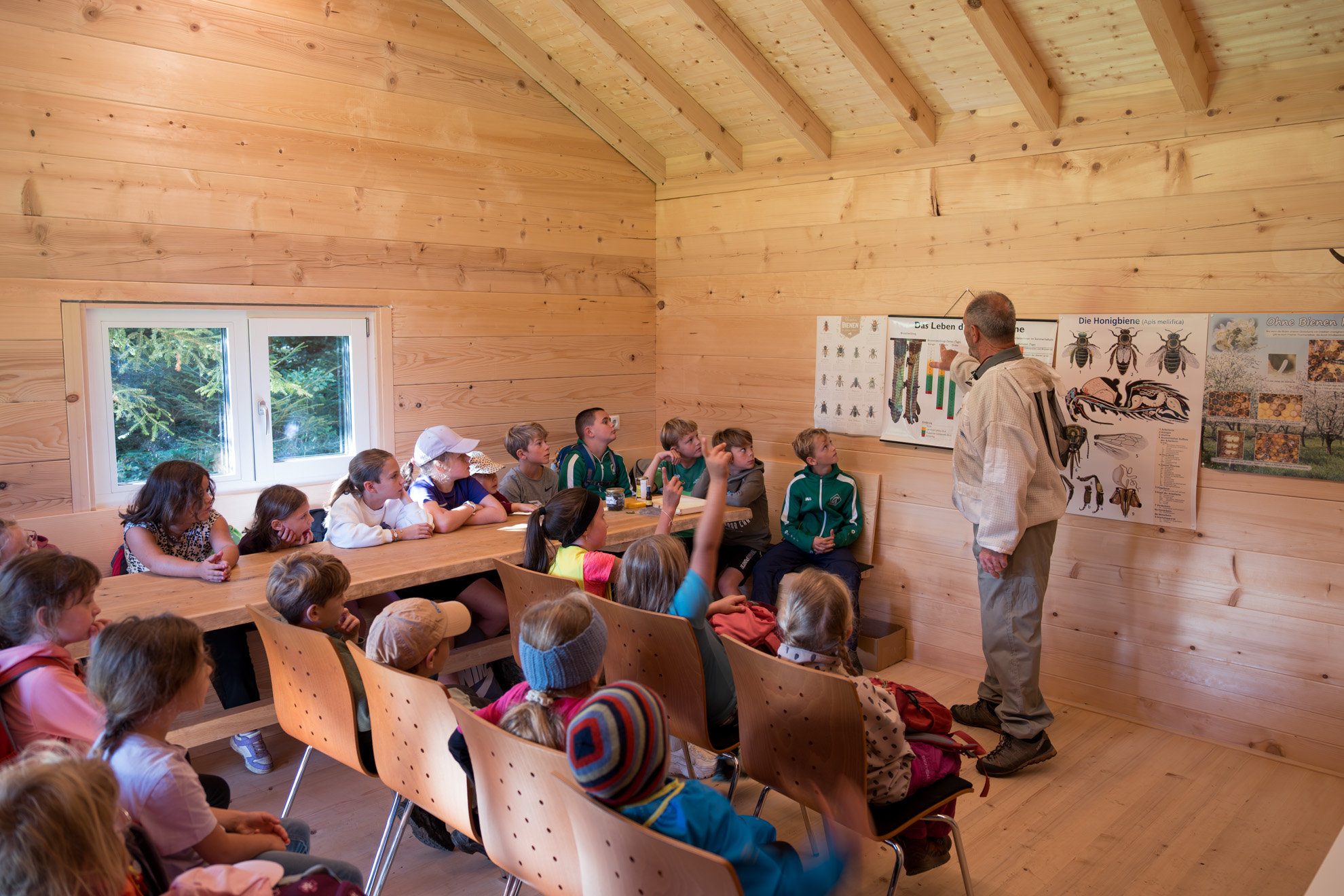 Kinder sitzen in einem Klassenraum aus Holz und hören einem älteren Mann zu, der eine Präsentation hält. Der Raum hat helle Holzverkleidungen an Wänden, Decke und Boden und wirkt rustikal und gemütlich. Der Mann steht vor einer Wand mit Lehrpostern, die Tiere, Spuren und ökologische Informationen zeigen. Die Kinder sitzen teils an einem Holztisch, teils in Stuhlreihen, und wirken aufmerksam. Ein Fenster auf der linken Seite gibt den Blick frei auf grüne Bäume, was auf eine Lage im Wald hinweist.