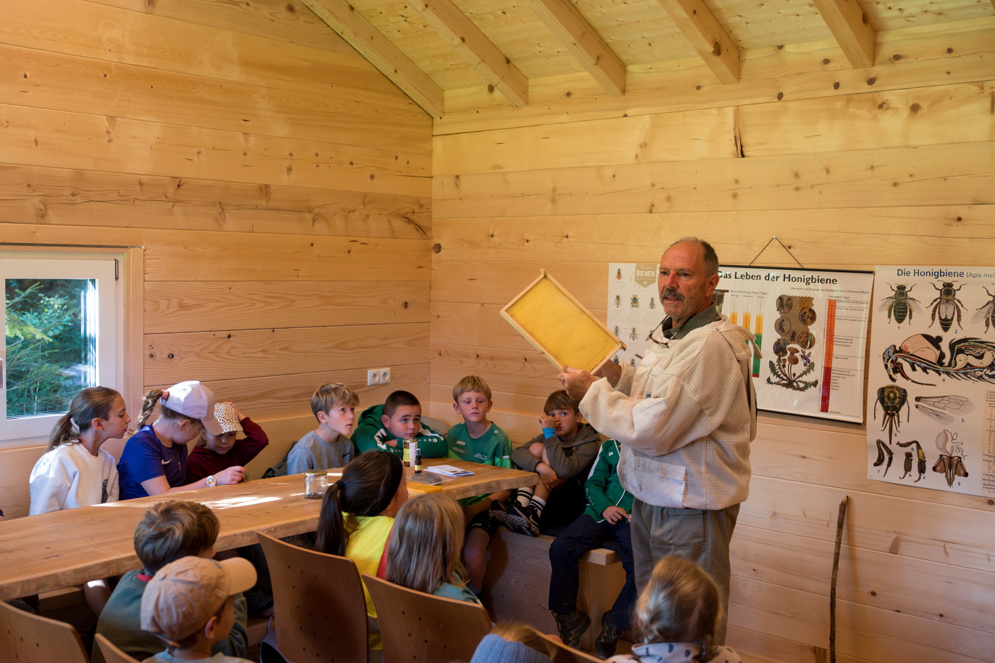 Kinder sitzen in einem Klassenraum innerhalb einer Holzhütte und hören einem männlichen Erwachsenen zu, der einen rechteckigen Holzrahmen – vermutlich ein Imkerrähmchen – in den Händen hält und etwas über Bienen erklärt. Die Wände bestehen aus hellem Holz, und an der Wand hinter dem Vortragenden hängen Poster mit Diagrammen und Informationen über Honigbienen, darunter anatomische Darstellungen und Lebenszyklen. Die Kinder sitzen an Holztischen und wirken aufmerksam und interessiert.