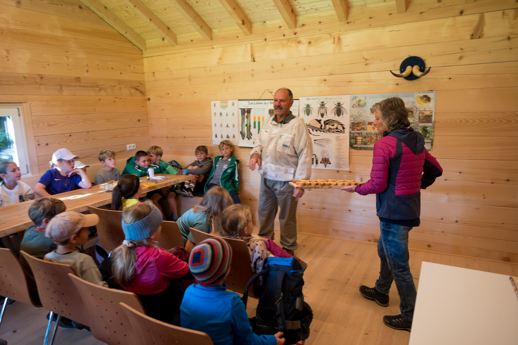 Kinder sitzen in einem Klassenraum innerhalb einer Holzhütte und hören aufmerksam zwei Erwachsenen zu. Eine Person trägt einen weißen Imkeranzug und scheint eine Präsentation über Bienen zu halten. Die andere Person hält ein Tablett mit Materialien oder Anschauungsobjekten. An der Wand hängen Poster mit verschiedenen Insekten, darunter auch Bienen, die das Thema des Unterrichts unterstützen. Die Kinder sitzen in Reihen auf Holzstühlen, und die warme Holzoptik des Raums schafft eine einladende Lernatmosphäre.